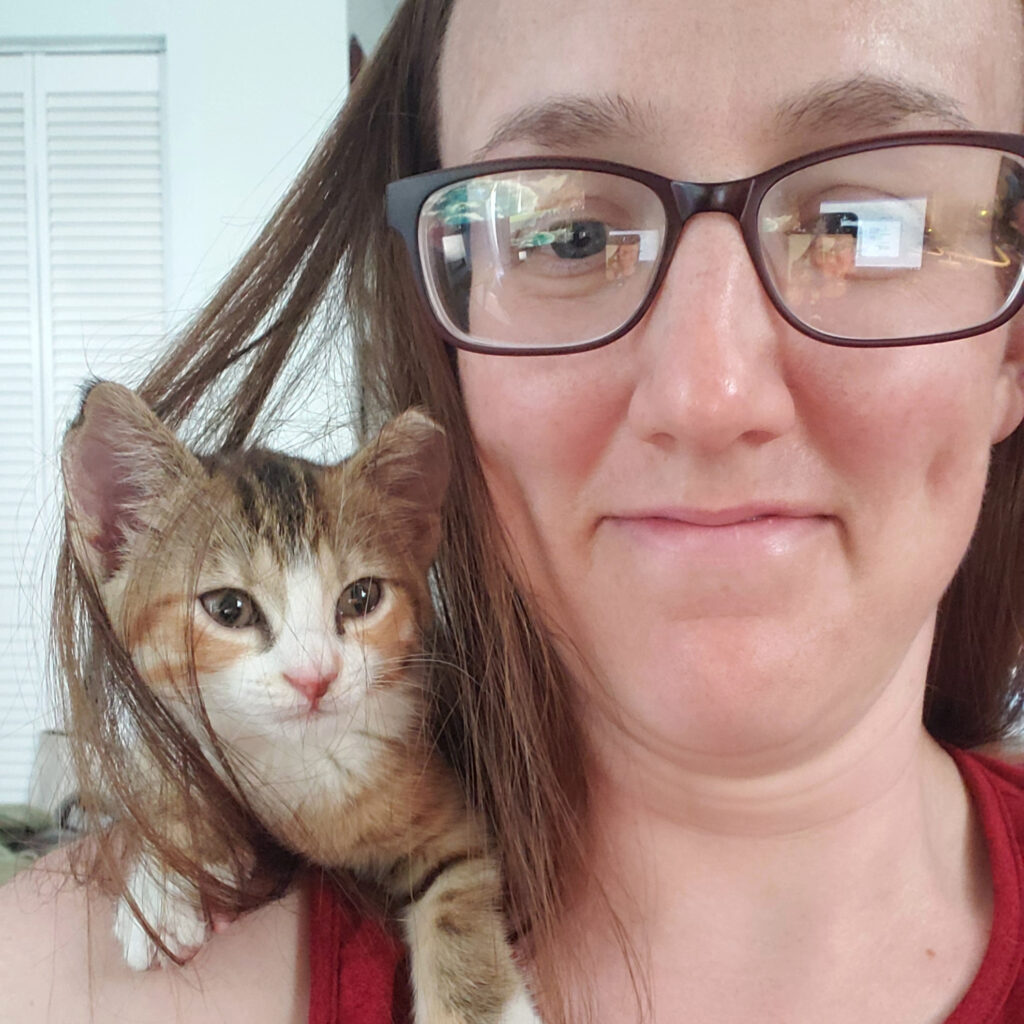 Woman with glasses with orange tabby kitten on shoulder.