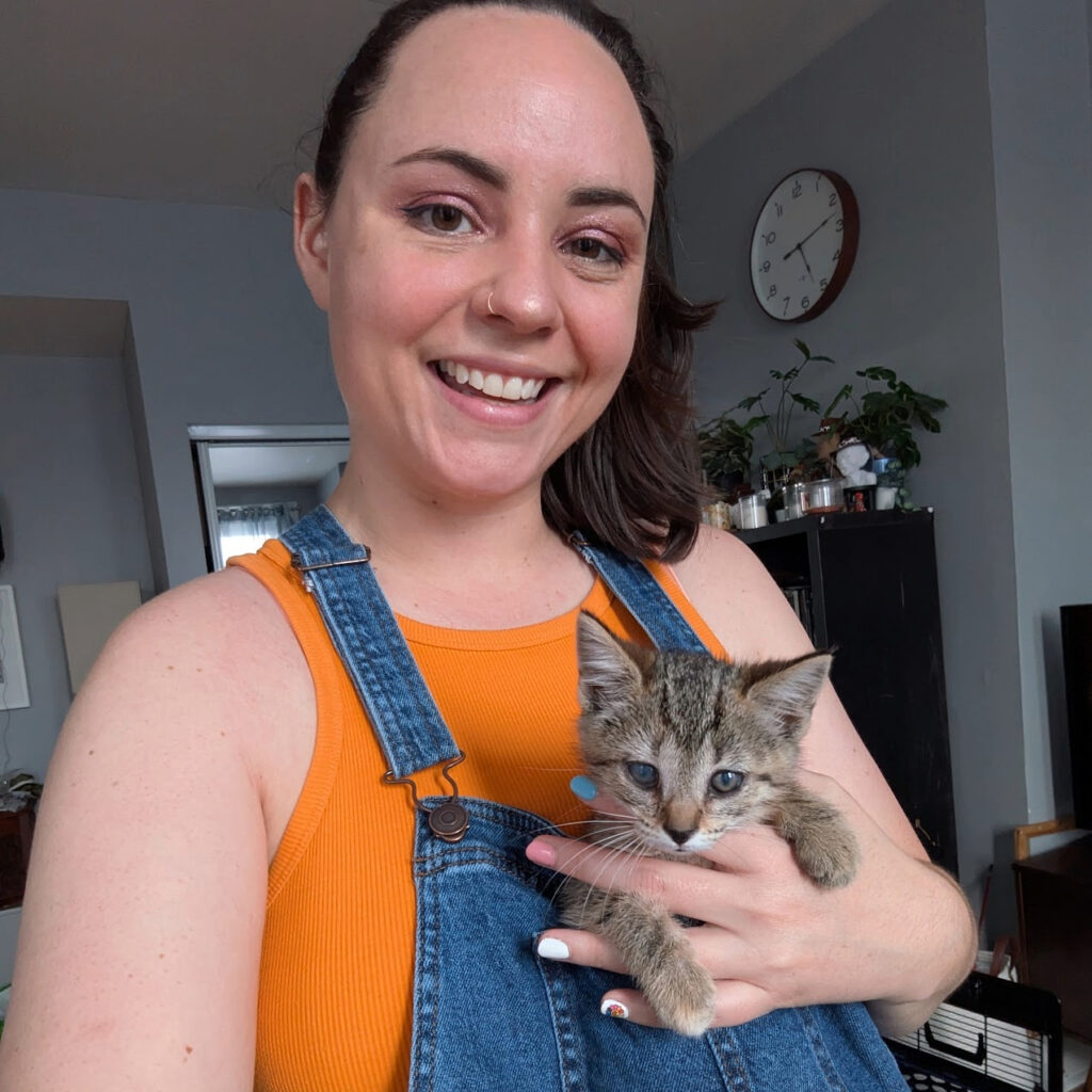 Woman with orange shirt and denim overalls holding a gray tabby kitten